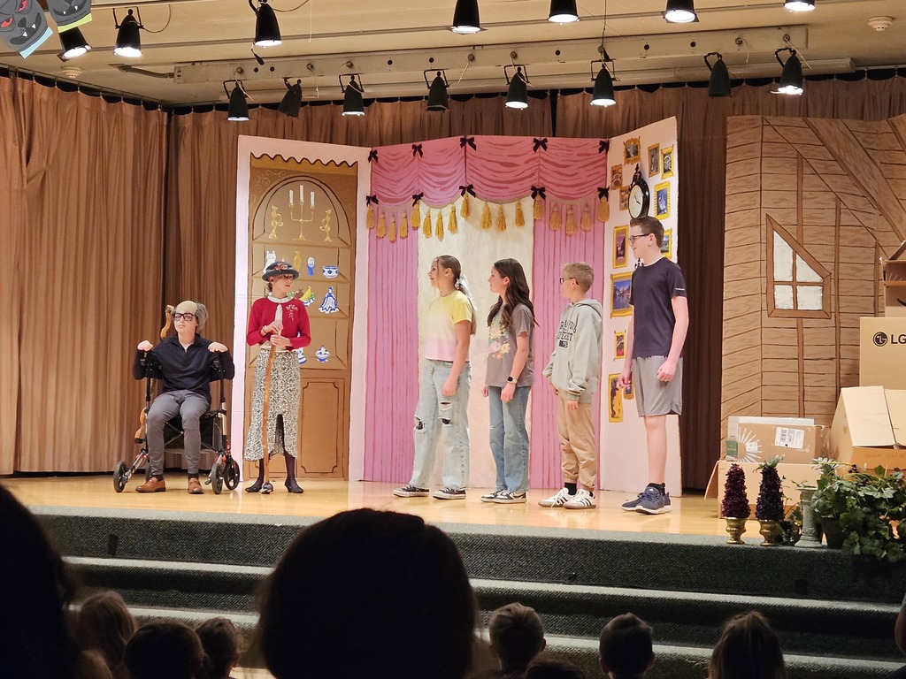 A scene from the play showing several students performing in front of a colorful set piece designed to look like an indoor room with pink curtains and a bookshelf.