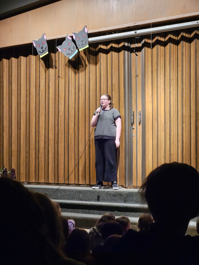 A young woman standing at a microphone on stage in front of a wooden-slatted backdrop, addressing the audience during a moment of recognition.