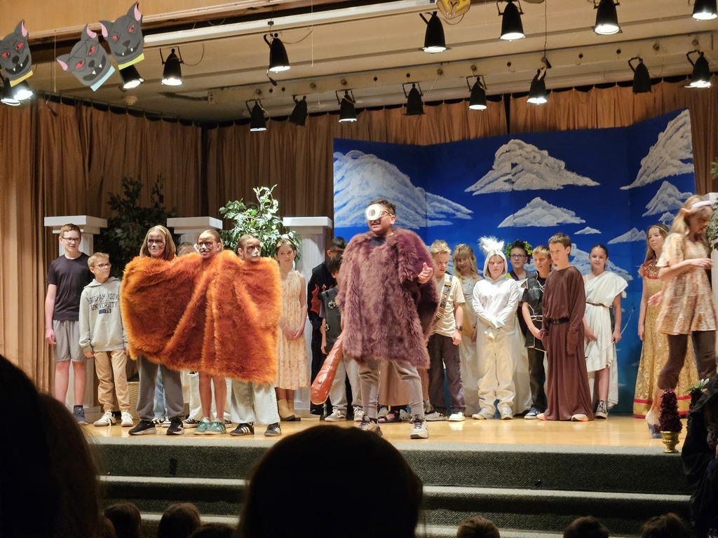 A standout character moment featuring a student in a large, shaggy purple monster costume with a single large eye on their forehead, standing center stage during the production.