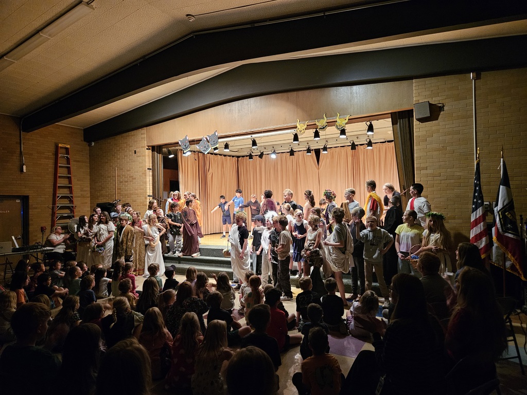 A wide view from the back of a packed school auditorium showing a large cast of 6th grade students in various Greek-themed costumes taking a bow on stage under bright theater lights.