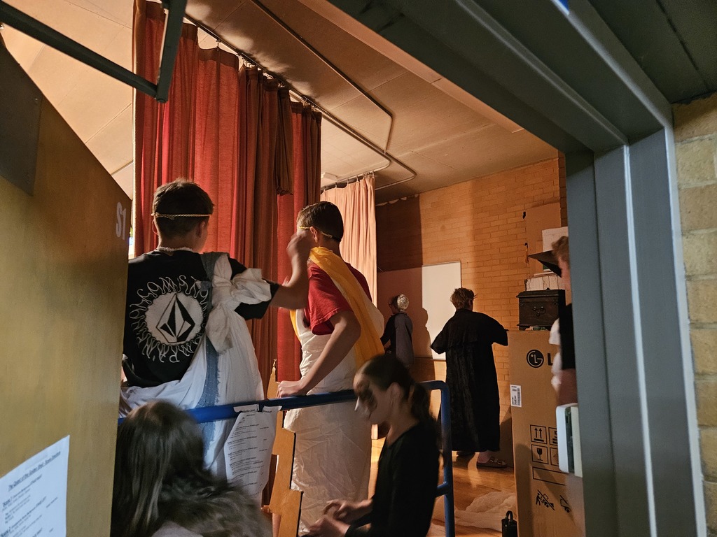 A candid behind-the-scenes shot from the wings of the stage, showing students in Greek tunics preparing for their entrance next to a large equipment box and red curtains.