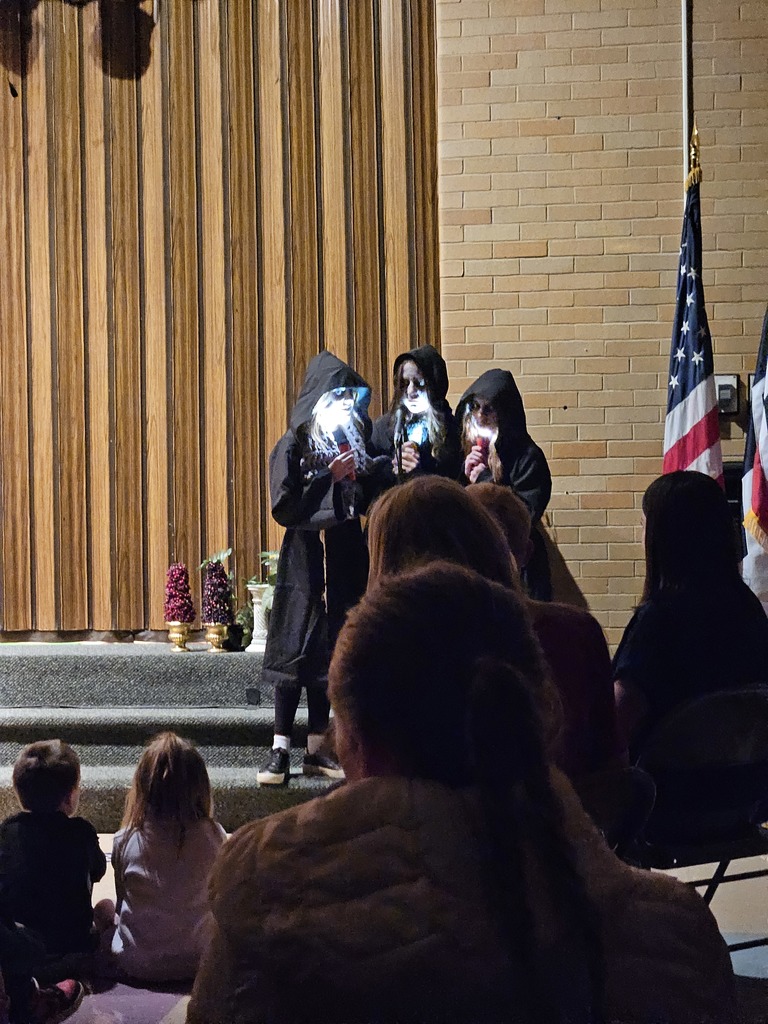 A moody, dramatic shot of three students dressed in dark hooded robes on a dimly lit stage, each holding a flashlight under their chin to create spooky shadows on their faces.