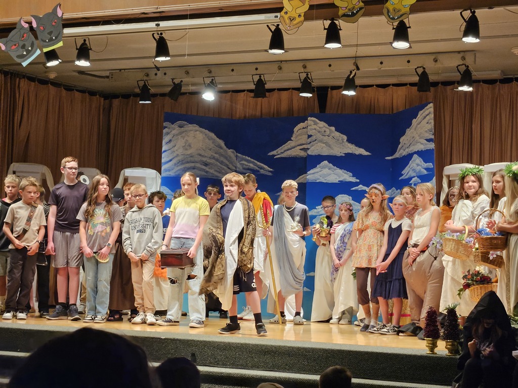 A wide view from the back of a packed school auditorium showing a large cast of 6th-grade students in various Greek-themed costumes taking a bow on stage under bright theater lights.
