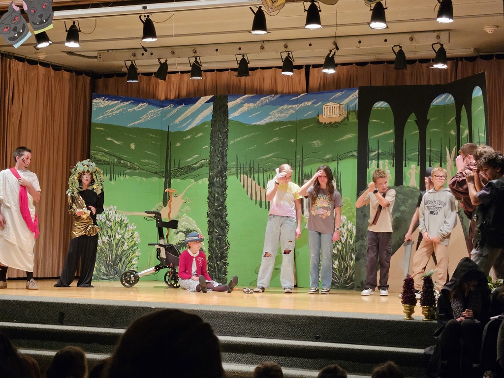 A stage scene featuring a student dressed as Medusa with a snake headpiece and gold costume, standing in front of a vibrant hand-painted backdrop of a Greek landscape with rolling green hills.