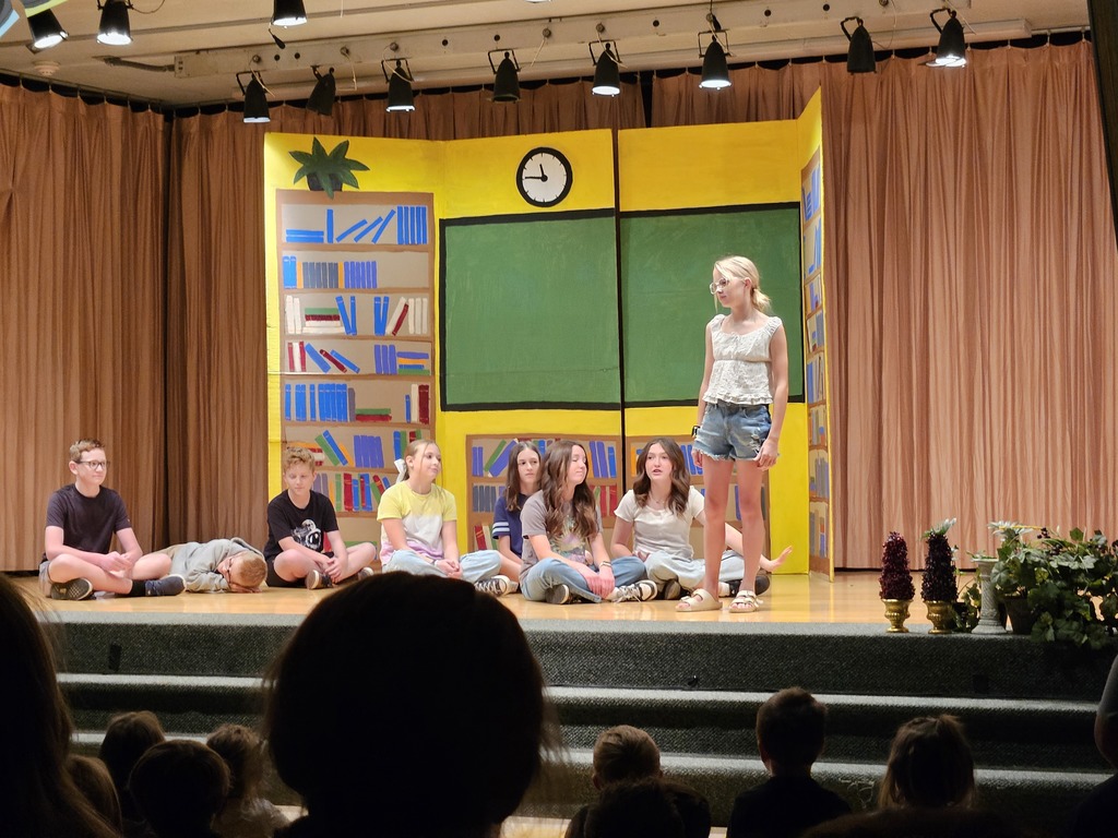 Students sitting on the stage floor in a semi-circle during a scene set in a classroom, featuring a large yellow and green set piece made to look like a chalkboard and library shelves.