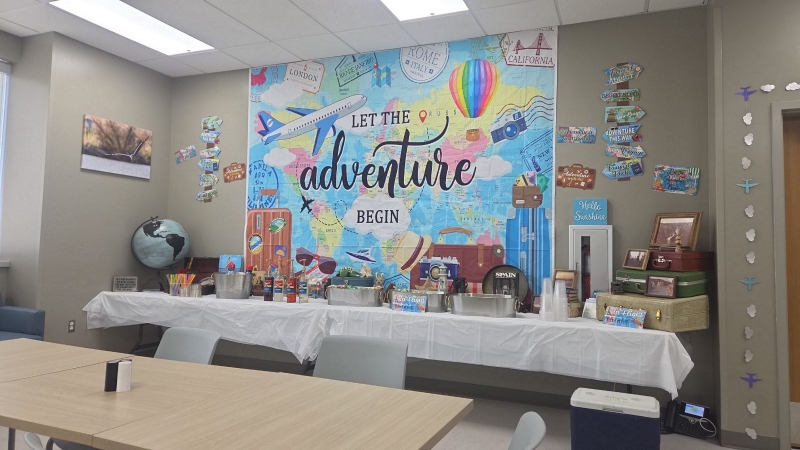 A wide shot of a community or break room decorated for a travel-themed event. A long table with a white cloth is positioned in front of a massive world map backdrop featuring the phrase "LET THE adventure BEGIN". On the table are drink tubs, syrups, and travel decor like a globe and a book titled "SPAIN".