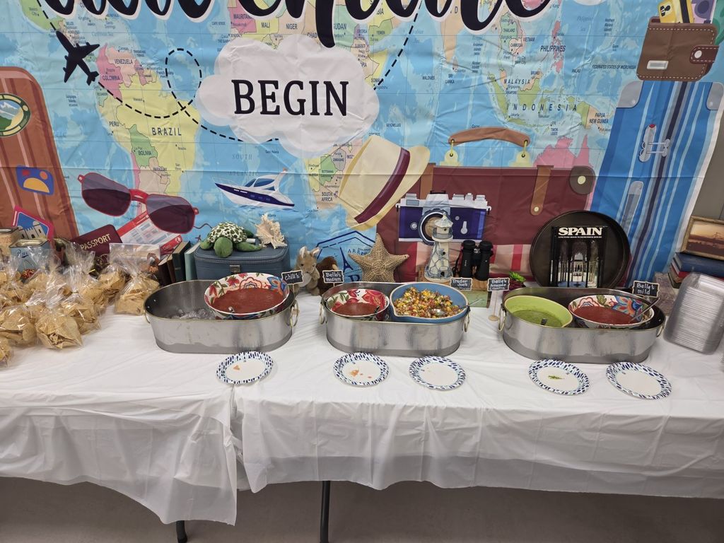 A high-angle shot of a salsa bar set up on a white table. Three large silver tubs hold various bowls of salsa and dips. Small chalkboard signs identify the flavors: "Chili's", "Bella's Medium", "Trader Joe's", "Bella's Avocado Cilantro", and "Bella's Mild". Bags of tortilla chips are visible on the left. The backdrop behind the table shows a world map with the word "BEGIN" in a cloud and travel graphics including sunglasses, a passport, a camera, and a book titled "SPAIN".