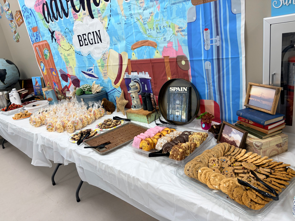 A close-up view of a long table covered in a white tablecloth, featuring an extensive spread of desserts and snacks. Items include bags of kettle corn, plates of brownies, a tray of pink and yellow macarons and chocolate-dipped cookies, and a large clear tray filled with chocolate chip and sugar cookies. In the background, a travel-themed backdrop features a world map and a large suitcase graphic. A black decorative plate rests on a stand with the word "SPAIN" and an image of Moorish architecture. The backdrop also features a cloud with the word "BEGIN" and a sign in the corner that says "Sunshine"