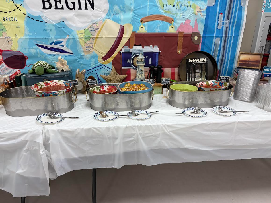 A close-up of a salsa station on a white-clothed table. Metal serving tubs contain colorful bowls of dip, including a corn salsa and a green avocado salsa. Small chalkboard labels read "Chili's", "Bella's Medium", "Trader Joe's", "Bella's Avocado Cilantro", and "Bella's Mild". Several small patterned paper plates with silver spoons are placed in front of the tubs.