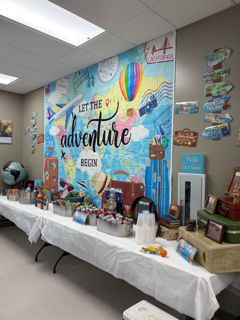 An indoor event space showing a long table prepared for a "Travel" theme. The table holds various "In-Flight Drinks," including sodas in metal tubs and flavored syrups. A large wall backdrop displays a world map with the text "LET THE adventure BEGIN", along with illustrations of a hot air balloon, a plane, and locations like "ROME ITALY", "LONDON", and "CALIFORNIA". Stacked vintage suitcases and directional signs such as "TRAVEL ADDICT", "DESSERT ISLAND", "Pack your Bags", and "PASSPORT Photos" decorate the right side. A small blue sign reads "Hello Sunshine".
