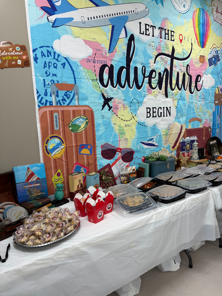 A buffet table set against a large travel-themed backdrop that reads, "LET THE adventure BEGIN". On the left, a large silver platter is piled high with individually wrapped fortune cookies. Next to them are several red Panda Express takeout boxes. The rest of the table is lined with black plastic catering containers with clear lids. The backdrop features a world map, an airplane, a suitcase with stickers, and the words "UNITED STATES", "BRAZIL", and "PHILIPPINES". A small sign on the wall to the left says "Adventure with me".