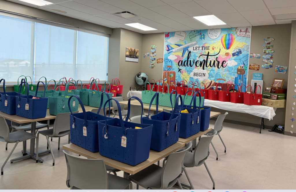 A large room filled with several rows of colorful rubber tote bags (Bogg Bags) in shades of blue, teal, and red, sitting on wooden tables and the buffet counter. Each bag contains snacks and has a gift tag attached that says, "THANKS FOR BEING AN Adventure". In the background, the travel-themed wall mural reads "LET THE adventure BEGIN". The room has large windows with closed blinds and gray chairs.