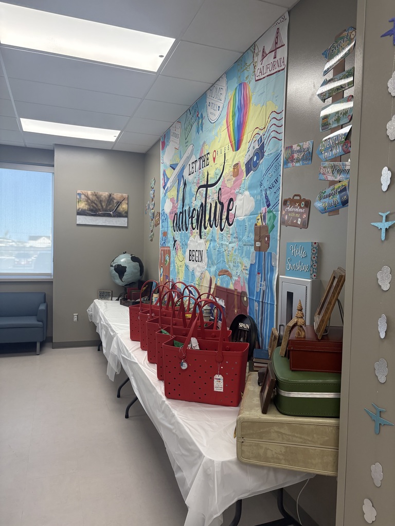 A side-angle view of a decorated room. Red rubber tote bags are lined up on a white-covered table in front of a travel backdrop that says "LET THE adventure BEGIN". To the right, a vertical stack of travel-themed signs includes "TRAVEL ADDICT", "Pack your Bags", and "ADVENTURE THIS WAY". A stack of vintage green and tan suitcases serves as a decorative base for framed photos and a blue sign that says "Hello Sunshine".