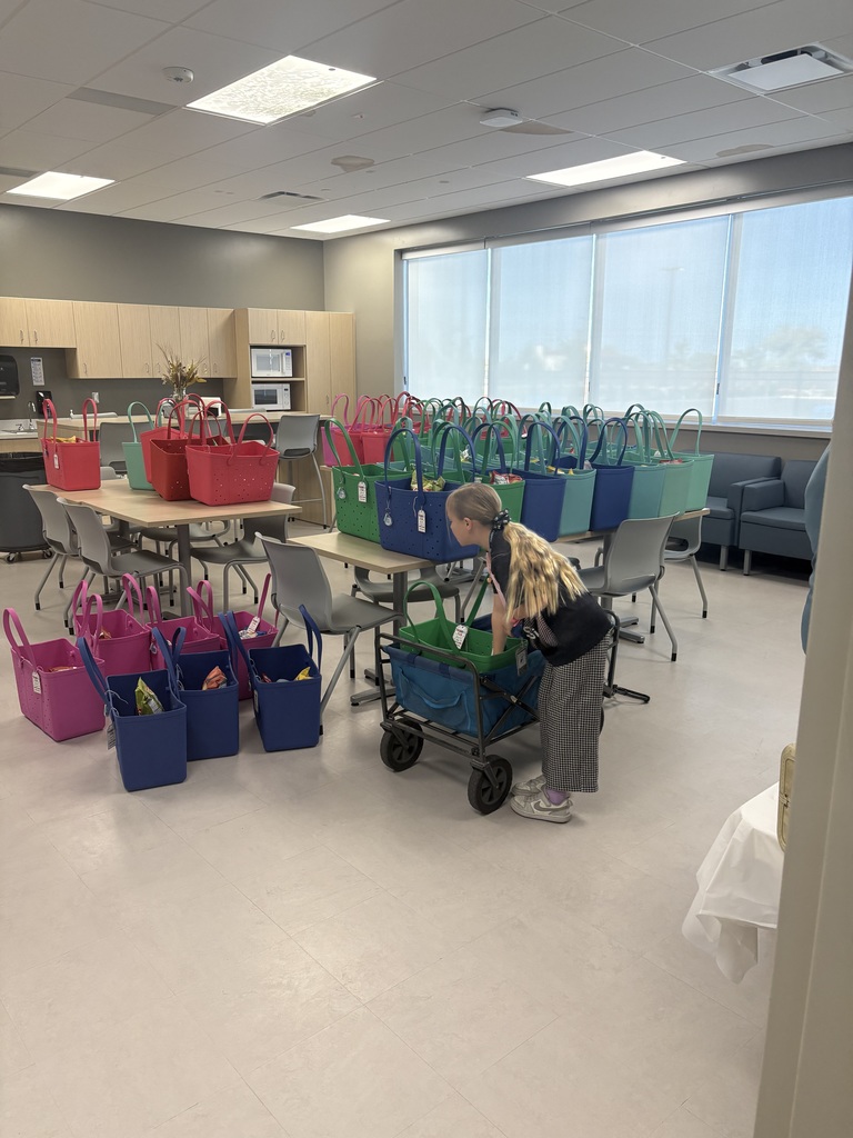 A young girl with long blonde hair in a ponytail is leaning over a blue rolling wagon in a large room. The room is filled with dozens of colorful rubber tote bags in pink, blue, teal, and green, arranged on top of and underneath several wooden tables. The bags appear to be prepared as gifts or party favors. In the background, there is a kitchen area with light wood cabinets and a microwave.