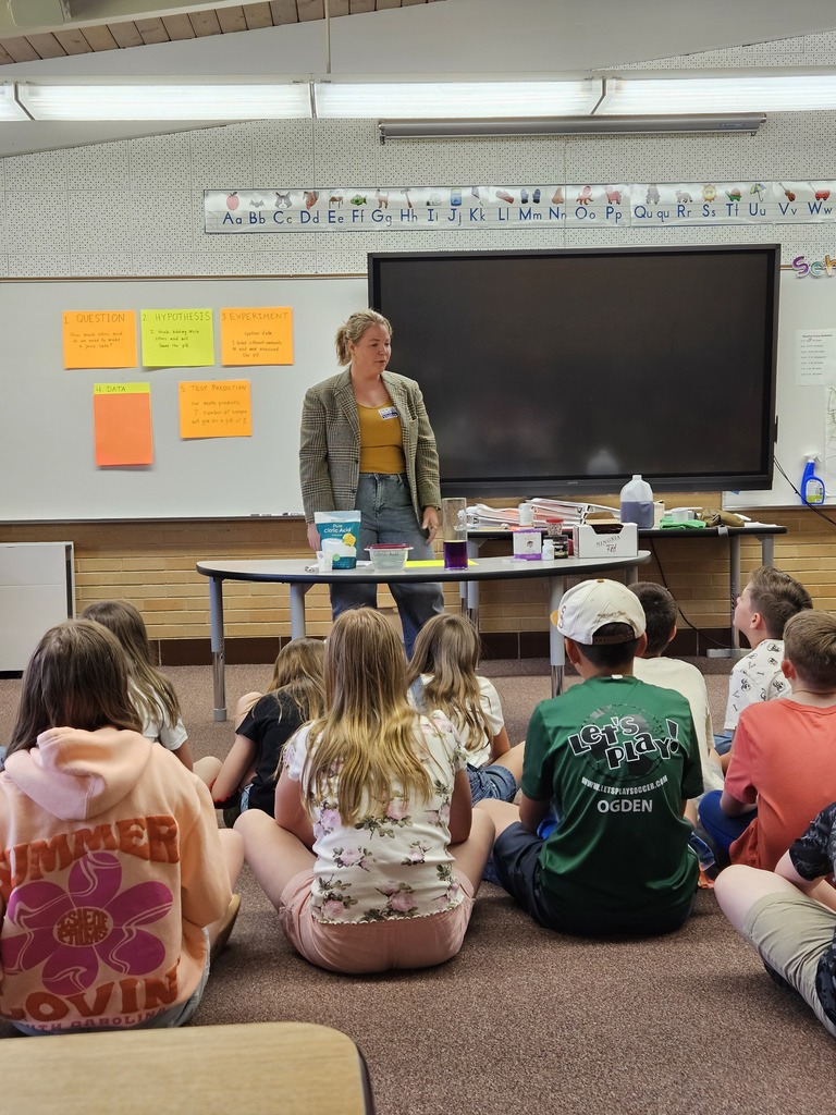 A presenter in a blazer stands behind a table with science experiments and "Hypothesis" posters on the wall.