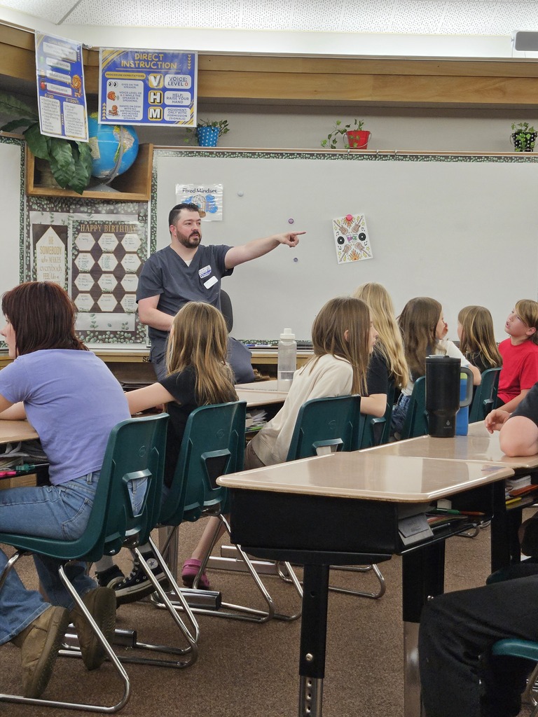 A male presenter in blue scrubs points across the classroom while students watch attentively.
