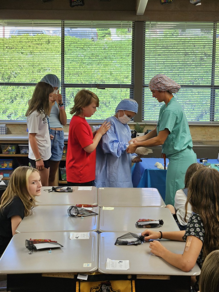 A female presenter in green surgical scrubs and a patterned floral scrub cap helps an elementary student put on white latex gloves. The student is already wearing a blue surgical gown and mask. Other students stand nearby or sit at their desks, watching the demonstration intently in a bright classroom with large windows in the background.