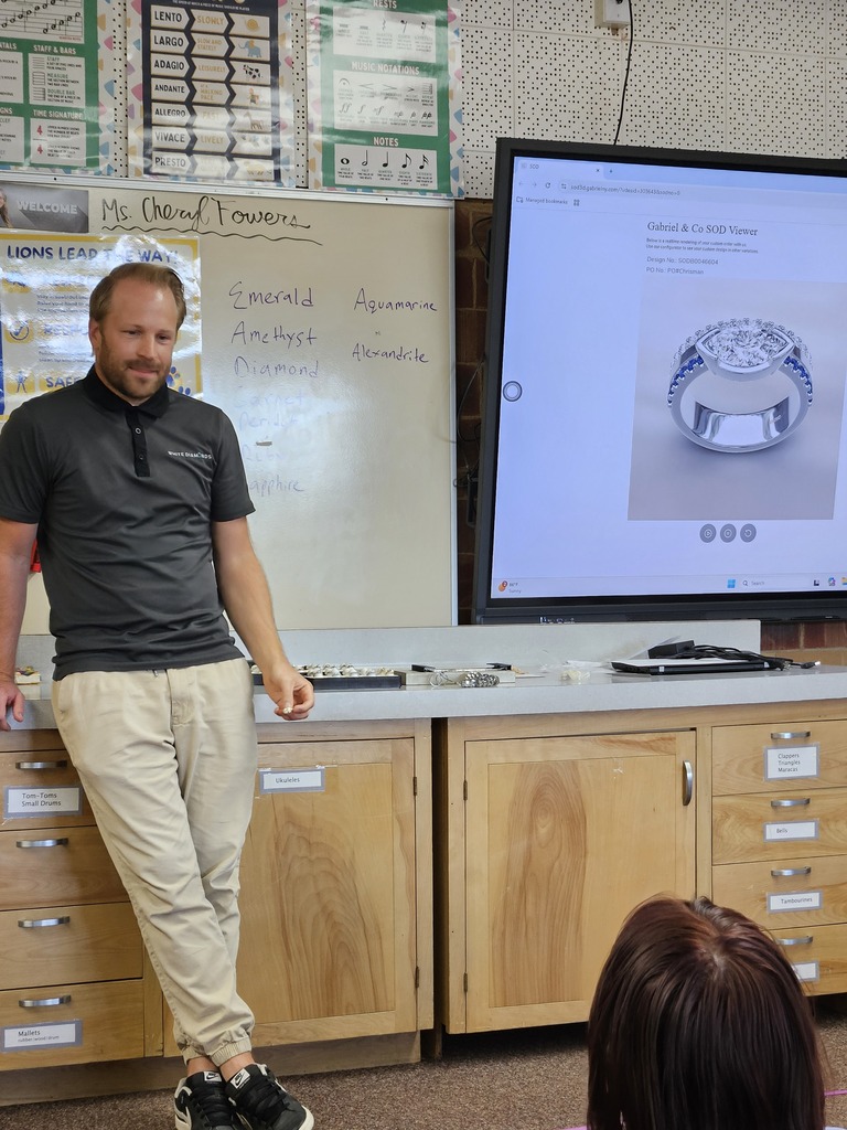 A male presenter in a polo shirt leans against a counter near a screen showing a 3D jewelry design of a diamond ring.