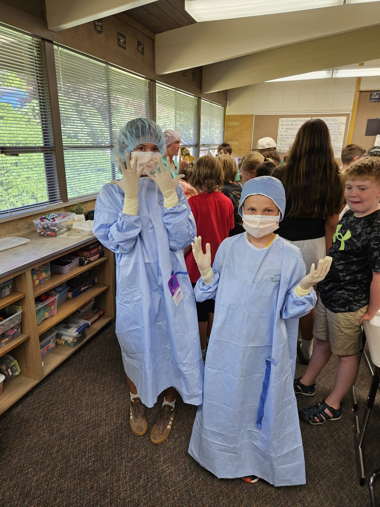 Two students dressed in full blue surgical gowns, masks, and gloves pose for the camera.
