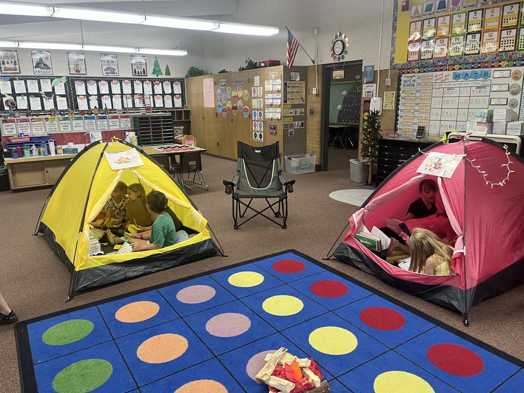  Multiple colorful tents (red, green, orange, and blue) are lined up in a classroom with various camping chairs and forest-themed decorations, creating a "tent city" for students.
