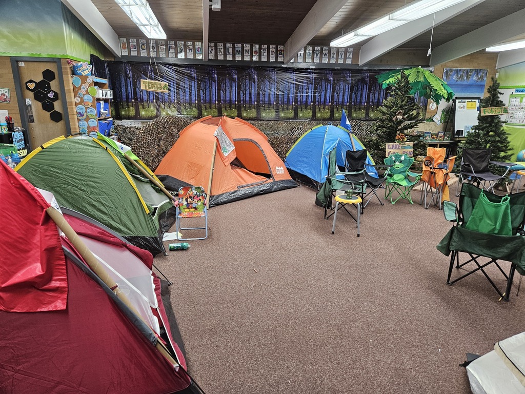 A wide view of a classroom transformed into a campsite with yellow and pink tents, camping chairs, and a pretend campfire made of logs and tissue paper on a colorful dot rug.