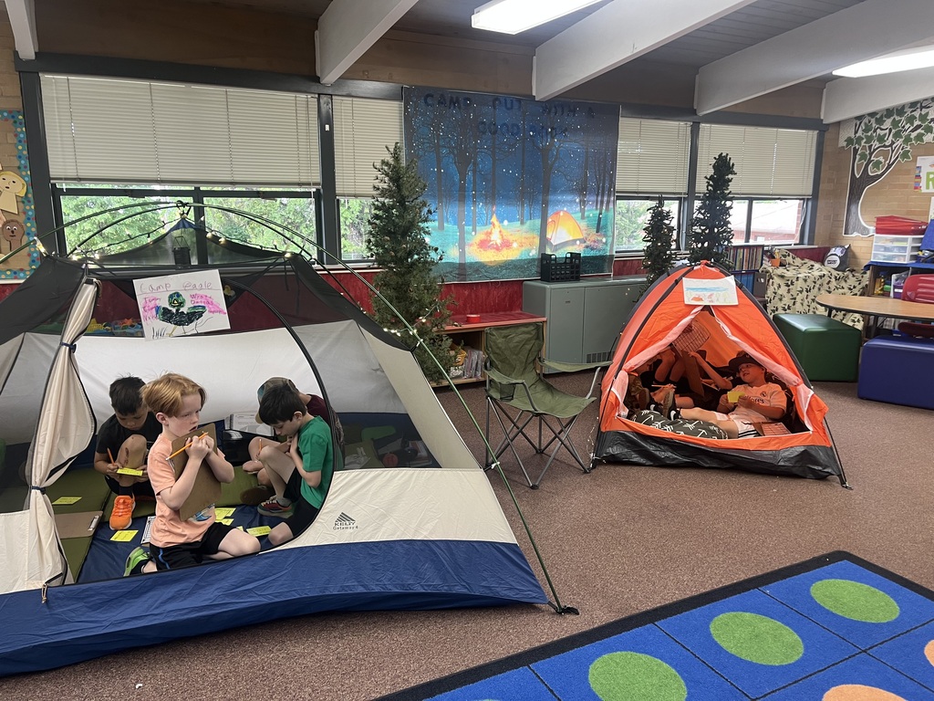 Students sit inside large camping tents set up in a classroom, using clipboards and pencils to work on assignments. A forest backdrop with a campfire hangs on the wall behind them.
