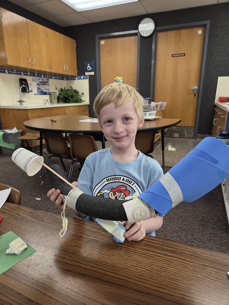 A young boy in a light blue t-shirt smiling while holding a creative "invention" made from a black foam noodle, blue paper, white cups, and masking tape.