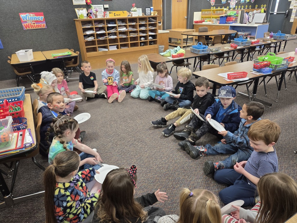A large group of kindergarten students sitting in a circle on a grey classroom carpet. Most students are holding white paper plates and looking toward the center of the circle during a group lesson.