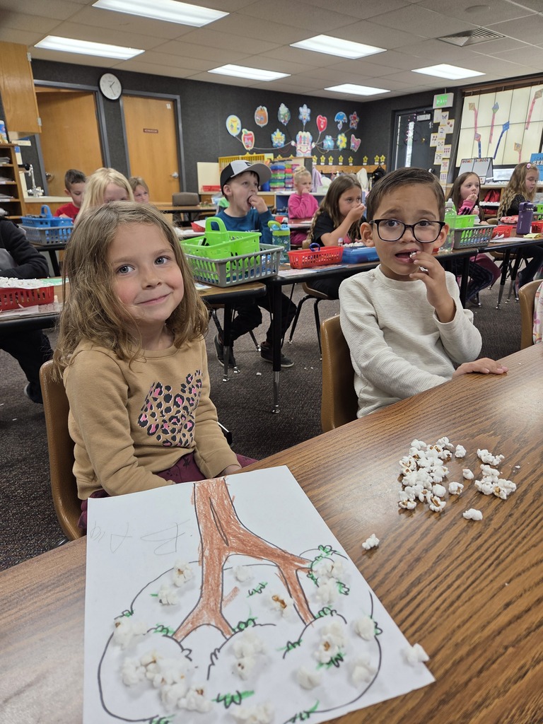 A young girl and boy sitting at a classroom desk. In front of them is a piece of art showing a brown tree trunk with real pieces of popcorn glued onto the branches to look like white blossoms