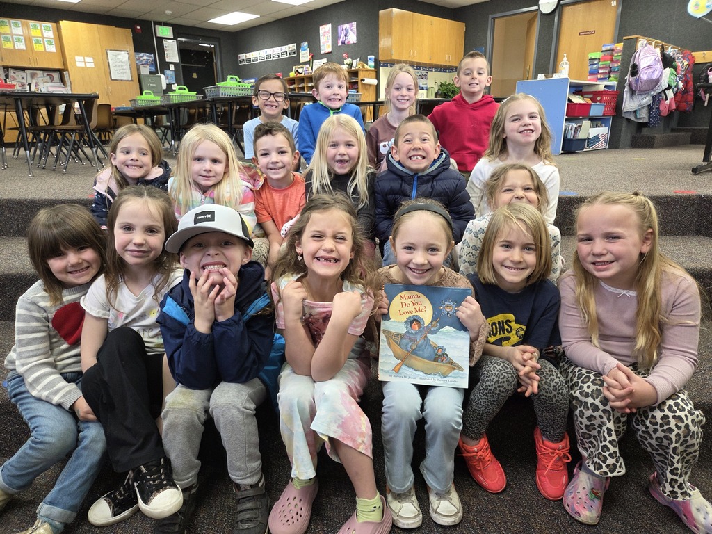 A group of about 18 smiling kindergarten students sitting on carpeted stairs in a classroom. A student in the front row is holding up the children's book "Mama, Do You Love Me?" for the camera.