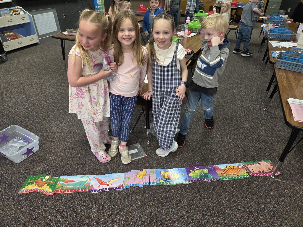 Four kindergarten students standing proudly behind a long row of completed picture puzzles laid out on the floor. The puzzles are numbered 1 through 10 and feature colorful illustrations of animals and insects.