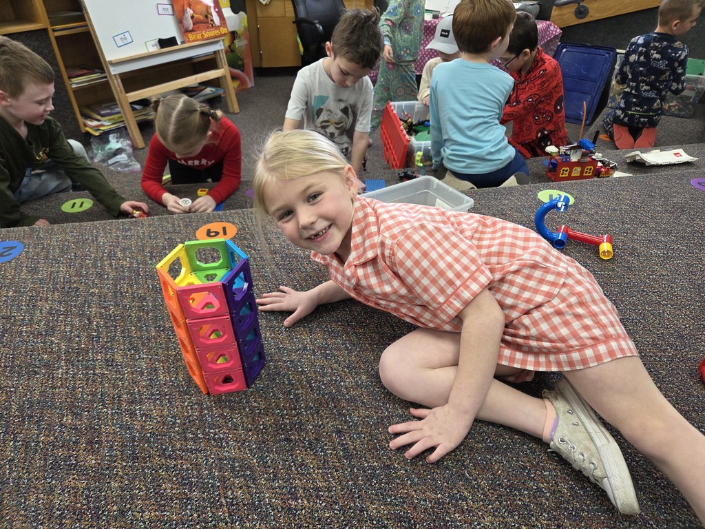 A young girl with blonde hair in a pink checkered dress smiles while lying on the floor next to a tall, colorful tower she built using geometric magnetic tiles.