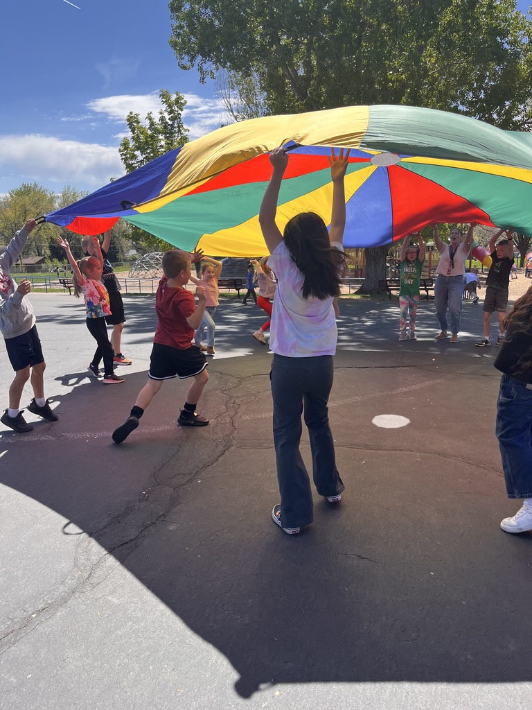 Students and a teacher stand in a circle on the playground, holding the edges of a large red, yellow, blue, and green parachute. They are lifting it together to create a giant "mushroom" shape under the bright afternoon sun.