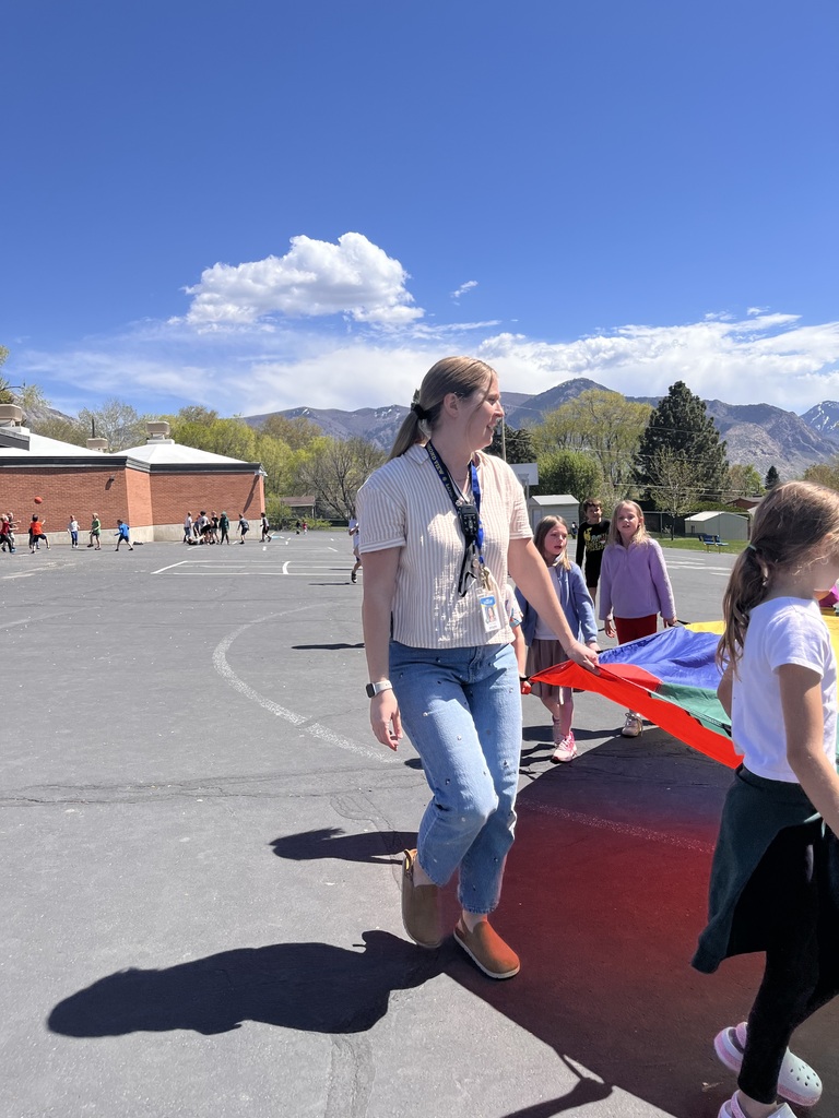 A female teacher wearing a striped shirt and jeans leads a group of students across a sunlit asphalt playground. They are carrying a folded, multi-colored parachute toward an open area, with mountains visible in the background under a blue sky.