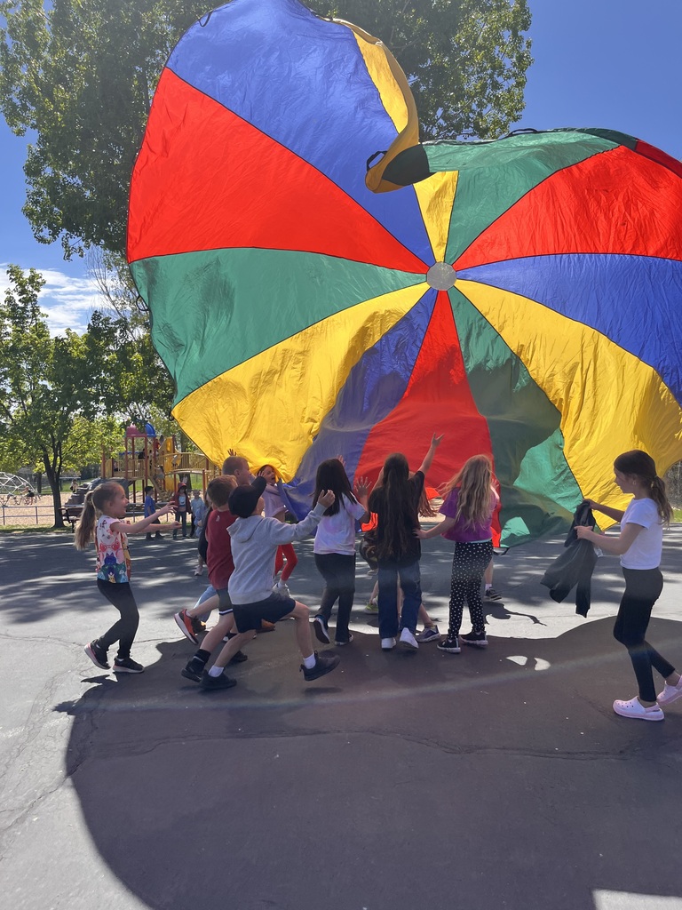 A group of elementary students stands under a large, colorful parachute that has been tossed high into the air. The bright sun shines through the fabric, creating a glowing effect as the children reach upward.