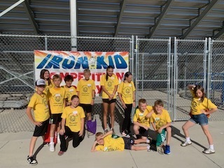 A group of students in yellow Iron Kid shirts posing in front of a colorful banner that reads "IRON KID COMPETITION" located at an outdoor athletic field with metal bleachers.