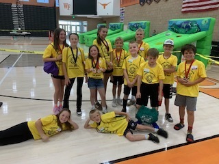 A smaller group of ten students in yellow Iron Kid shirts posing in a school gymnasium. In the background, there are large green inflatable obstacle course games.