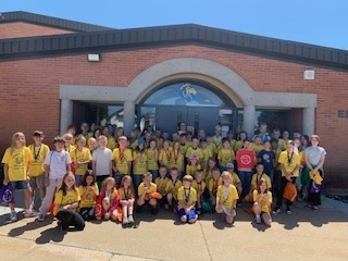 A large group of elementary students from Lomond View wearing matching yellow "Iron Kid" t-shirts, posing together in front of the brick school entrance under a clear blue sky.
