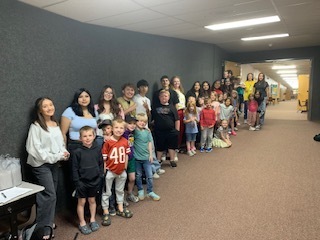 A large, diverse group of elementary school students and older student mentors or teachers standing in a long line against a dark gray wall in a school hallway. The group includes children of various ages smiling for the camera, with a brightly lit hallway extending into the background.