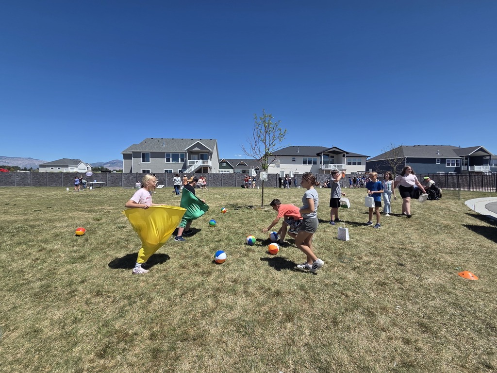 A group of children play a game on a wide, sunny grass field under a bright blue sky, with suburban houses and distant mountains in the background. In the foreground, one child runs while holding open a large yellow fabric sack, and another runs nearby with a green fabric sack. Colorful beach balls are scattered across the grass as other children watch and participate.