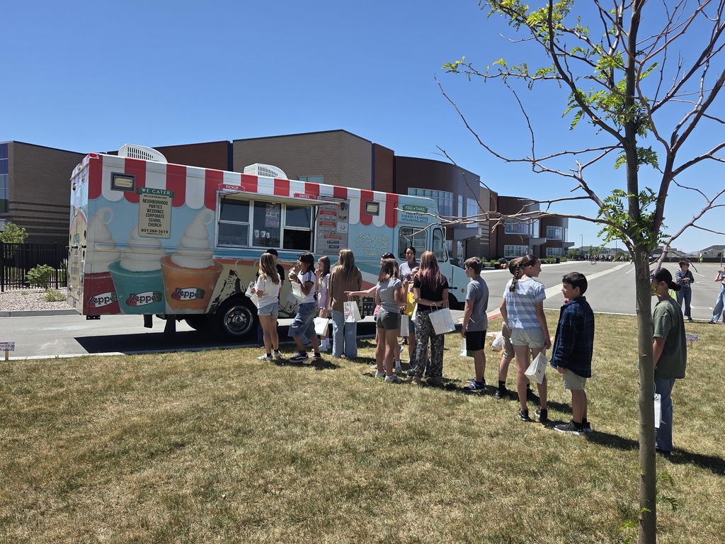 A long line of children stands on the grass waiting to order at a large, colorful Italian ice food truck parked outside a modern school building under a clear blue sky.
