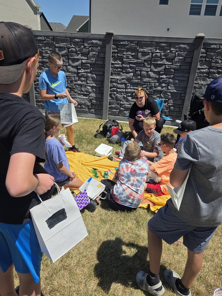A high-angle shot shows a group of kids gathered around a yellow blanket laid out on the grass next to a dark grey stone-patterned fence. An adult woman sits nearby watching them. The children are holding white paper gift bags, and one boy wearing a blue Nike shirt has a small white tag stuck to his forehead.