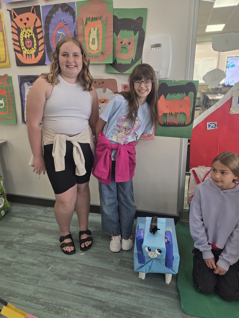 Two smiling young girls stand side-by-side next to a wall covered in colorful monster artwork, while a third girl sits on the floor to the right. In front of them on the floor is a blue boxy cardboard animal craft with black paper hair on top and a drawn-on face.
