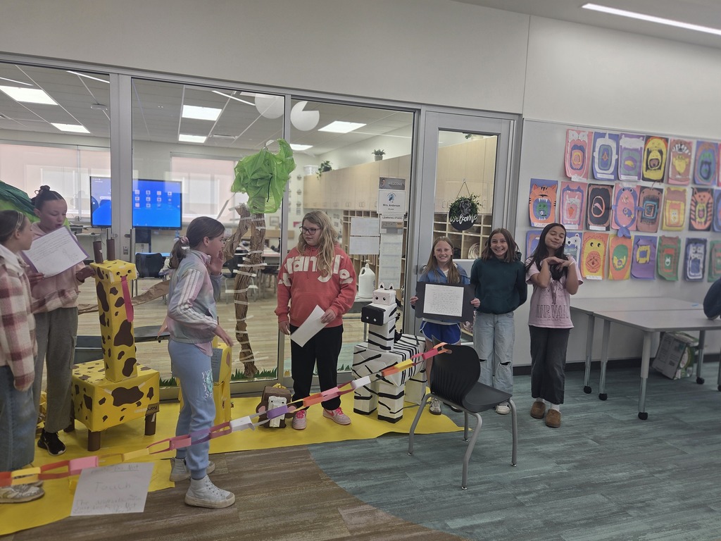 A group of seven elementary school students present their handmade cardboard animal projects inside a bright school hallway. On the left, a tall yellow giraffe with brown spots stands on a yellow paper mat. In the center, a student stands next to a white and black striped cardboard zebra. Paper chain link barriers separate the display from the walkway. The background features glass windows looking into a classroom and a wall covered in colorful student drawings of monster faces.