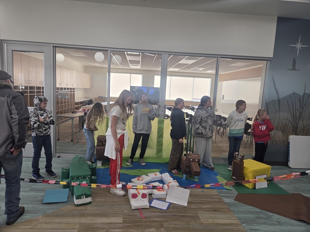 A group of students gather around an ecosystem display laid out on blue and green paper on a school hallway floor, representing an ocean or island habitat. The display features blocky cardboard animals, including a green crocodile, a white snake, and a brown hippo. A multi-colored paper chain barrier runs across the foreground, separating the viewers from the project.