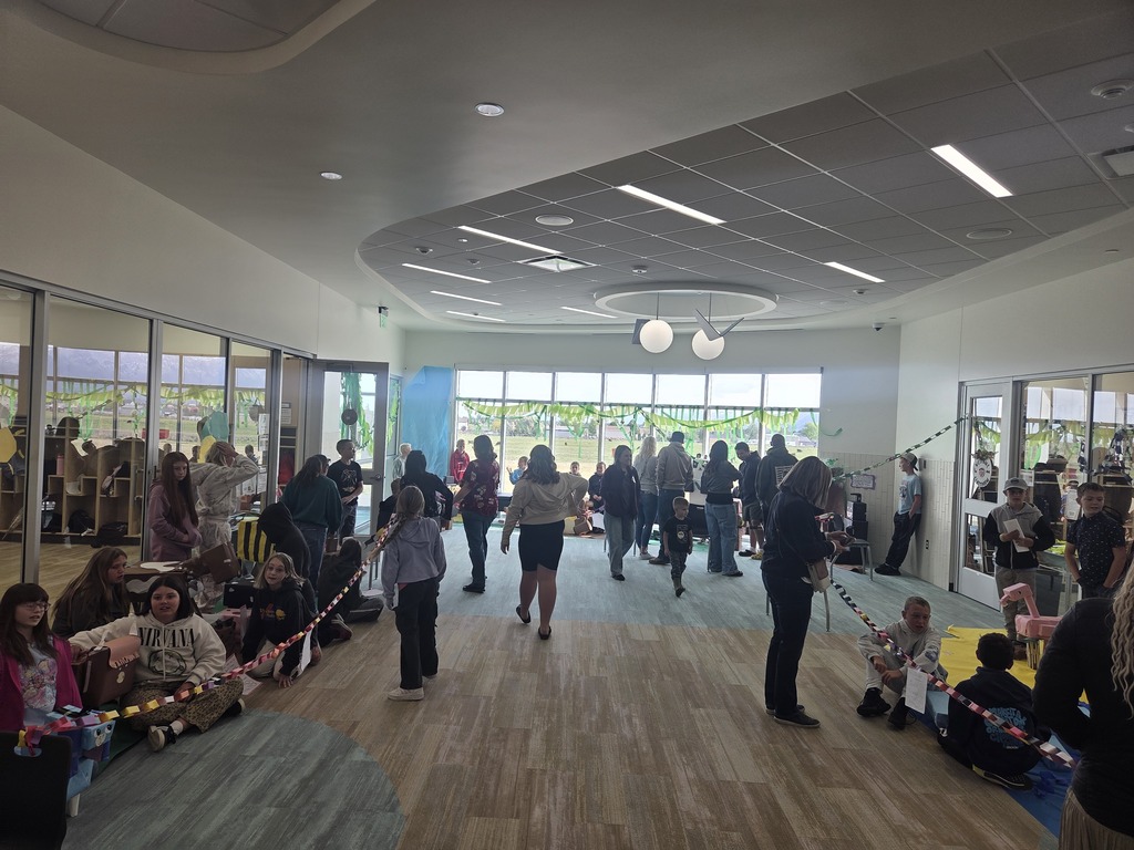 A bustling school hallway hosting a student art and science exhibition. A long white wall on the right is covered with rows of vibrant, colorful paintings of open-mouthed monsters. On the floor below the art, students sit next to blue paper displays representing water habitats with pink and blue cardboard animals. Paper chains stretch across the hallway to create viewing boundaries as parents and students walk through.
