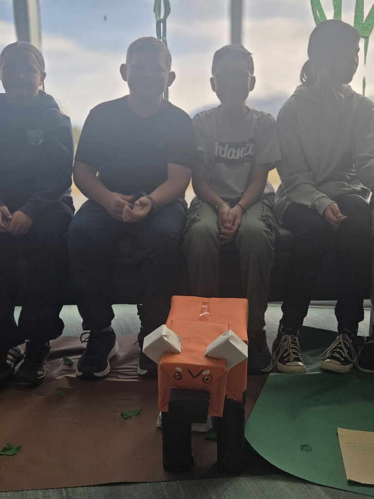 A close-up shot of a student-made, boxy orange cardboard fox or red panda sitting on the floor in front of four elementary school students who are sitting on a bench. The cardboard animal features white folded paper ears, a black block snout, and drawn-on eyes and nose. The students' faces are softly blurred by background light from a large window behind them.