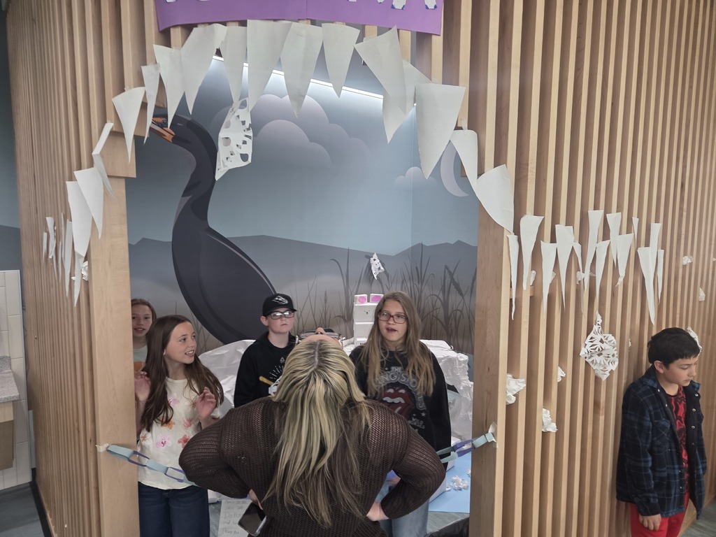 Several elementary school students stand inside a decorated alcove designed to look like a giant animal's open mouth, complete with large white paper triangles hanging down like sharp teeth. The background wall inside the alcove features a painted mural of a large dark bird, possibly a cormorant or heron, against a wetland landscape under a cloudy sky. An adult with blonde hair stands with her back to the camera, talking to the kids.