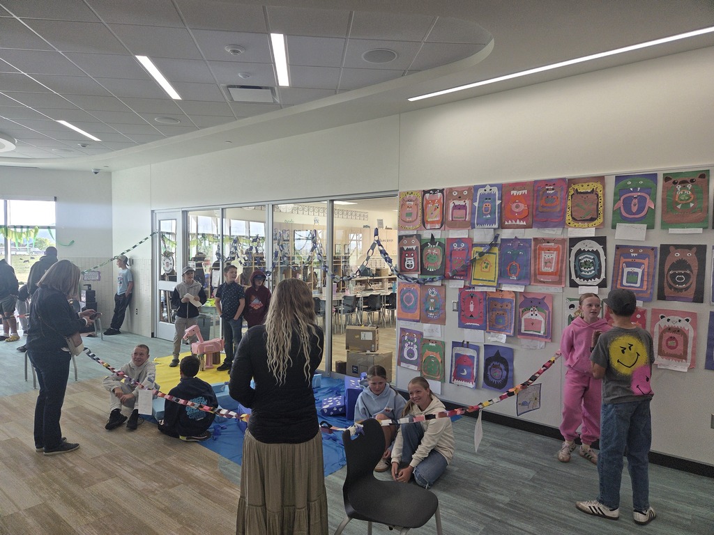 A wide view of an elementary school exhibition in a hallway outside classroom glass walls. Several students stand around various handmade boxy cardboard animals, including an orange fox, a yellow lion with a yarn mane, a yellow giraffe, and a striped zebra, all arranged on yellow paper sheets on the floor. A multi-colored paper chain link fence sections off the display area. Adults and students are seen interacting.