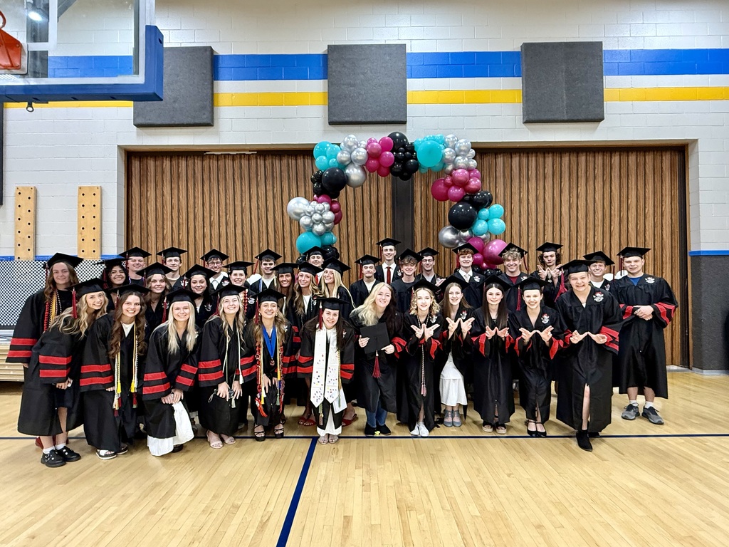 A large group of high school graduates in caps and gowns posing for a group photo in the Lomond View gym in front of a colorful balloon arch.